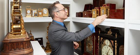 A man in a grey suit and glasses is facing left in a small room filled with shelves of antique clocks