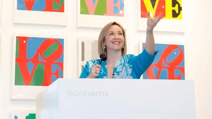 A woman in a blue top stands at a podium labelled Bonhams, holding a gavel and gesturing upwards. Colourful artworks spelling LOVE are displayed on the wall in the background.