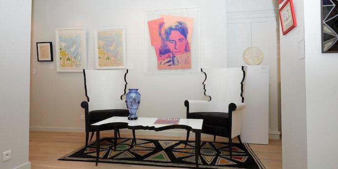 Two white armchairs and matching coffee table stand in a white room, surrounded by framed artworks on the walls and a patterned rug on the floor.