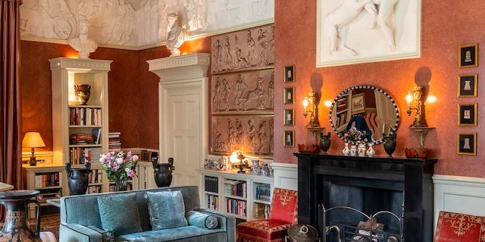 A room with orange walls is filled with objects, including marble carvings, bookshelves, a blue sofa on the left and a black fireplace on the right.