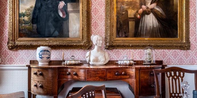 An antique wooden desk stands in front of a pink patterned wall, surrounded by large paintings, sculptures and other works of art.